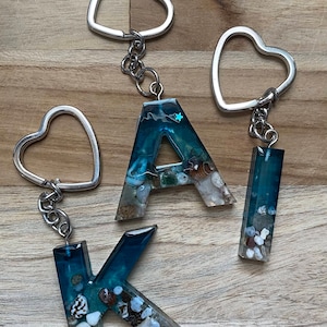 Top view of a customizable ocean-themed Resin Initial Keychain made with blue epoxy resin, in which tiny seashells and stones are present.