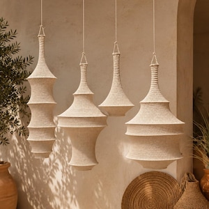 May include: Four off-white, hand-crocheted pendant light shades of varying shapes and sizes hang from thin cords. Each shade features a dense, textured weave with tiered, geometric silhouettes, suspended at different heights in a row.