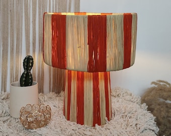 May include: A red and cream striped table lamp with a round base and a cylindrical shade. The lamp is sitting on a white fluffy rug.