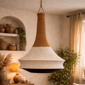 May include: A hanging, handcrafted crochet lamp with a brown top, white middle, and black bottom. The lamp is suspended from the ceiling in a cozy, well-lit living room. The room features a wooden coffee table, sofa, and decorative items.