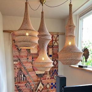 May include: Three hanging pendant lights with woven tan shades suspended from the ceiling. Each light features a tapered, multi-tiered silhouette with a visible internal bulb. The fixtures are positioned in front of a large, rectangular wall tapestry.