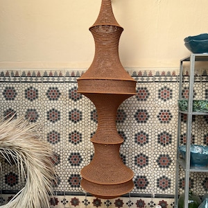 May include: A tall, vertically oriented hanging light fixture featuring a brown, crocheted fiber shade with a complex, multi-tiered hourglass silhouette. The structure consists of two wide, rounded sections connected by a narrower middle, tapering to a point.
