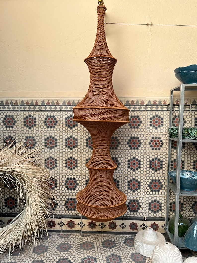 May include: A tall, vertically oriented hanging light fixture featuring a brown, crocheted fiber shade with a complex, multi-tiered hourglass silhouette. The structure consists of two wide, rounded sections connected by a narrower middle, tapering to a point.