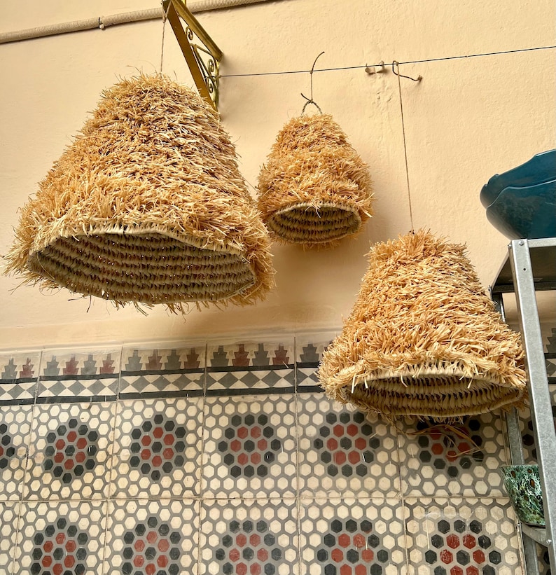 May include: Three hanging lampshades made of woven straw. The lampshades are cone-shaped with a woven base and a shaggy, textured exterior. The lamps are suspended from the ceiling by a wire. The background features a patterned tile wall.