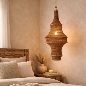 May include: A brown woven pendant light fixture with a tapered, multi-tiered silhouette suspended from a brass-colored chain. The light features a dense, intricate mesh pattern that allows a warm, soft glow to emit from the center bulb.