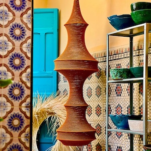 May include: A tall, orange, woven pendant light fixture with a symmetrical, multi-tiered hourglass shape. The structure features a fine, textured mesh pattern throughout, suspended from a metal bracket attached to the wall above.