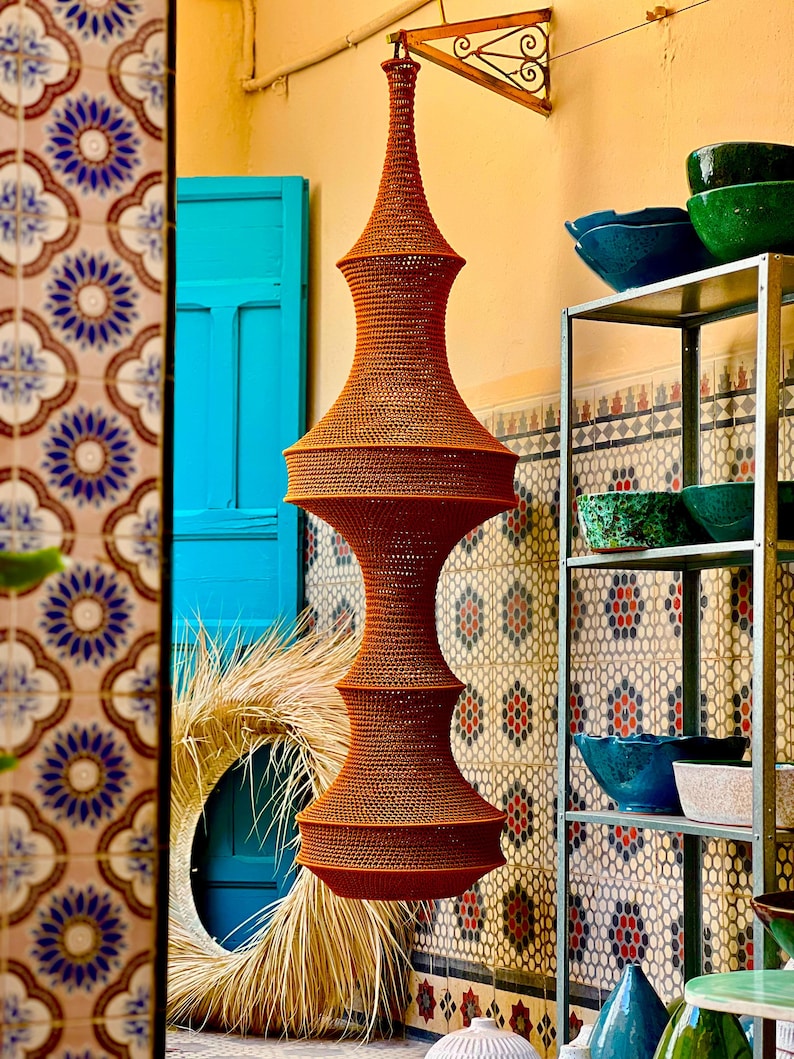 May include: A tall, orange, woven pendant light fixture with a symmetrical, multi-tiered hourglass shape. The structure features a fine, textured mesh pattern throughout, suspended from a metal bracket attached to the wall above.