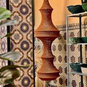May include: A tall, hanging pendant light fixture crafted from a rust-colored, woven material. The structure features a symmetrical, multi-tiered silhouette with a narrow top that expands into two distinct, wide, rounded sections.