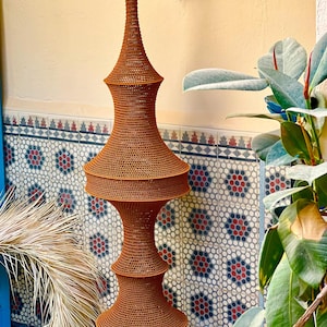 May include: A tall, rust-colored, woven hanging lamp fixture with a multi-tiered, hourglass-inspired silhouette. The structure features a fine, mesh-like texture throughout, suspended from a gold-toned metal wall bracket.