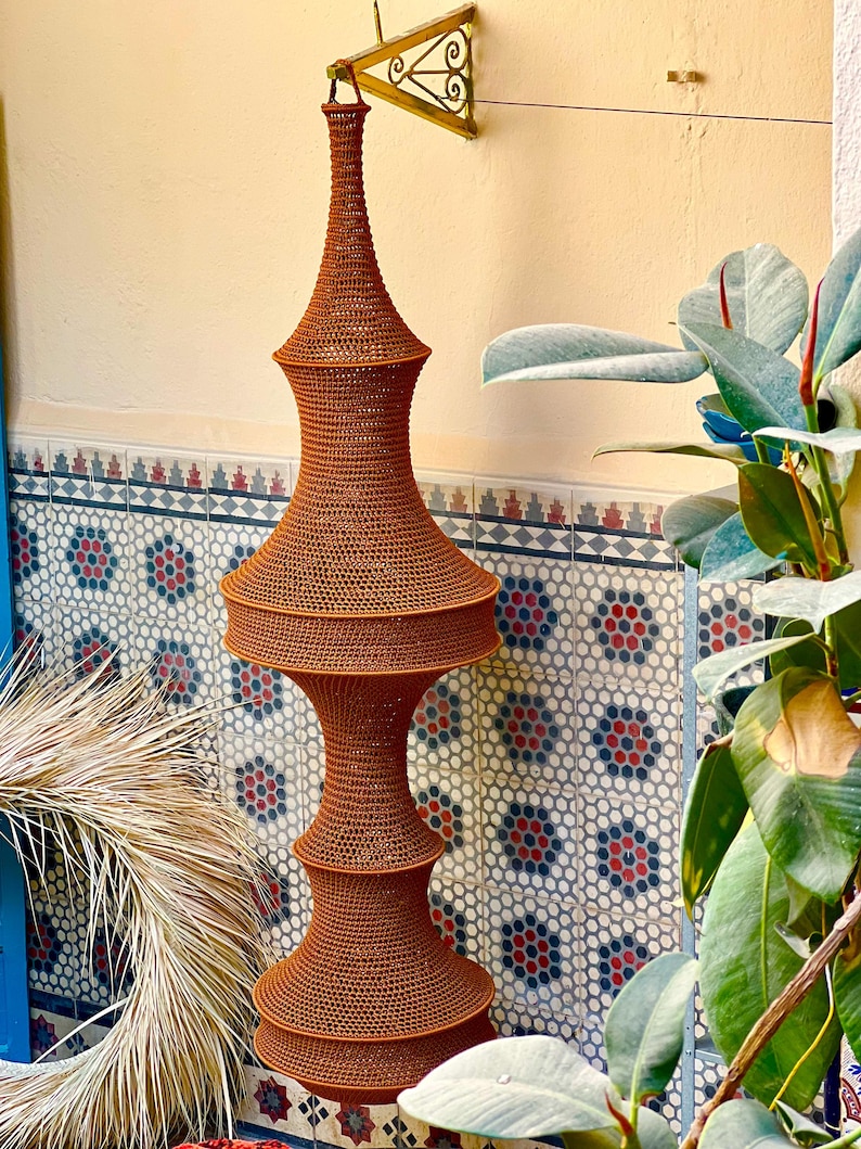 May include: A tall, rust-colored, woven hanging lamp fixture with a multi-tiered, hourglass-inspired silhouette. The structure features a fine, mesh-like texture throughout, suspended from a gold-toned metal wall bracket.