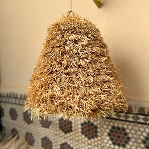 May include: A hanging lampshade with a conical shape, crafted from natural-colored, shaggy straw. The lampshade is suspended from a thin wire, casting a warm, inviting glow. The background features a patterned tile wall.