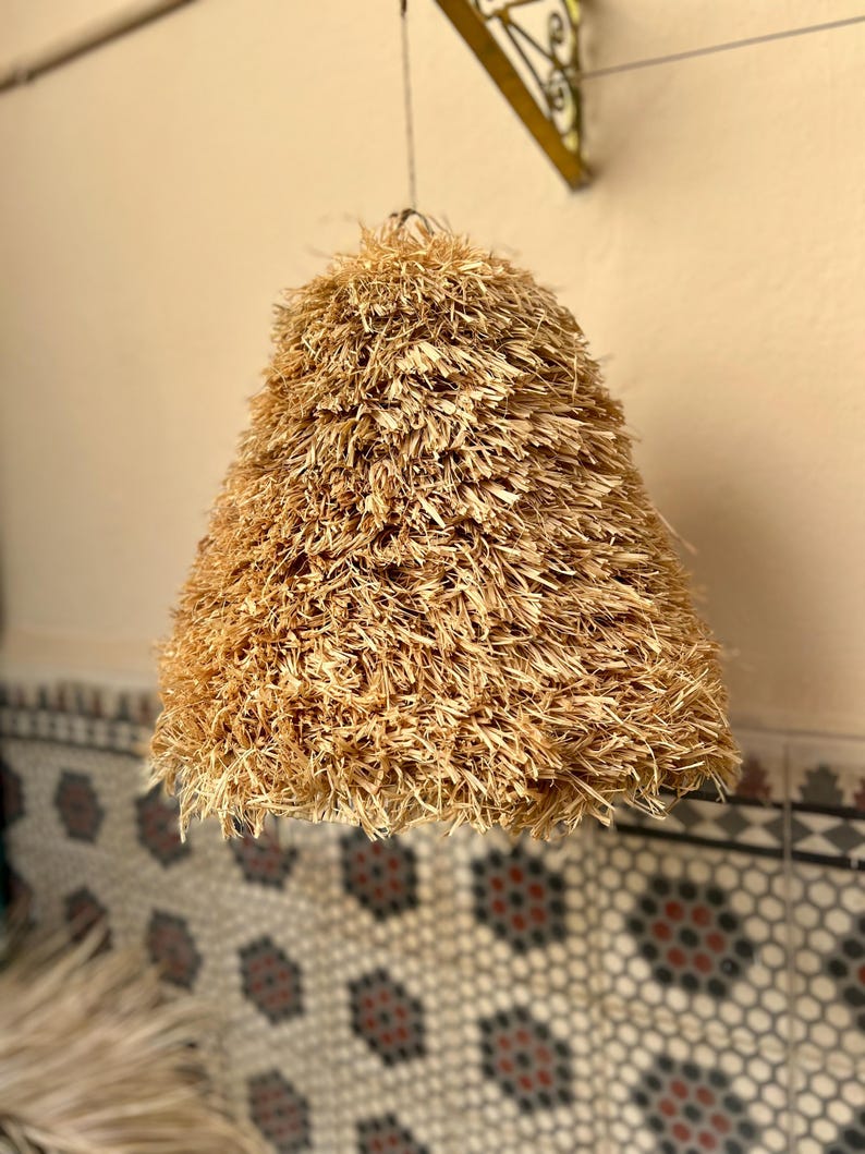 May include: A hanging lampshade with a conical shape, crafted from natural-colored, shaggy straw. The lampshade is suspended from a thin wire, casting a warm, inviting glow. The background features a patterned tile wall.