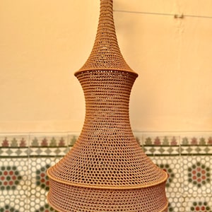 May include: A tall, brown, crocheted pendant light shade with a tapered, hourglass-like silhouette. The structure features a fine, open-weave mesh pattern throughout, with a wider, circular base section that narrows toward the top suspension point.