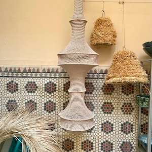 May include: A tall, beige, crocheted pendant light fixture with a multi-tiered, hourglass-like silhouette. The structure features a dense, intricate mesh pattern throughout, suspended from a single point at the top by a small metal loop.