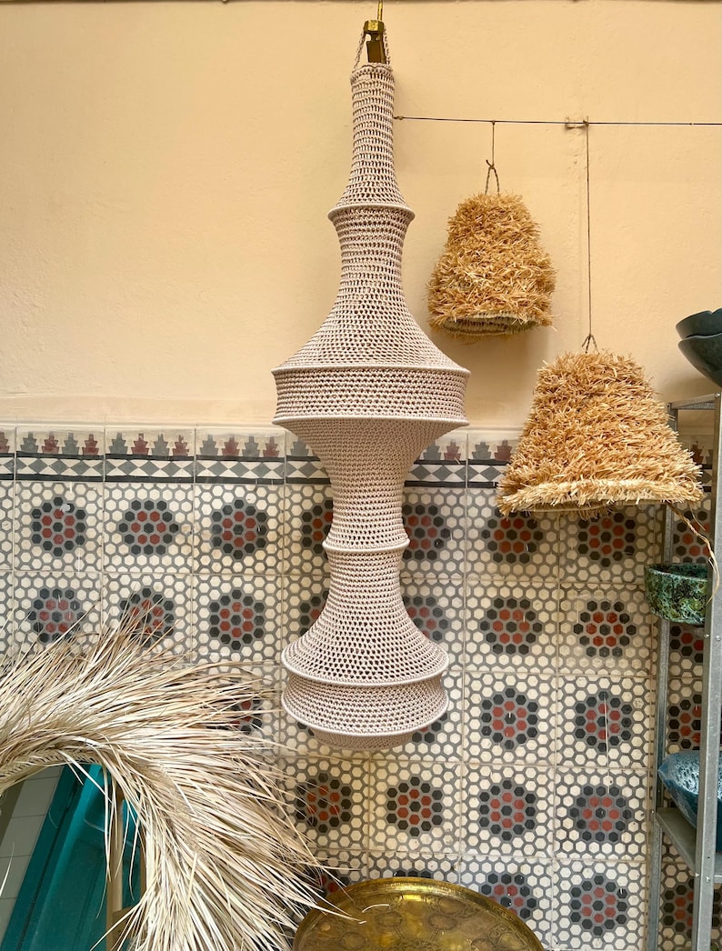 May include: A tall, beige, crocheted pendant light fixture with a multi-tiered, hourglass-like silhouette. The structure features a dense, intricate mesh pattern throughout, suspended from a single point at the top by a small metal loop.