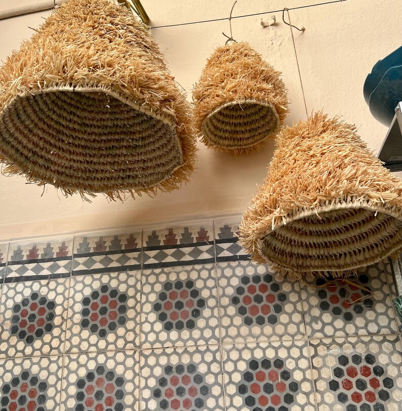 May include: Three hanging woven lampshades with a conical shape. The lampshades are made of natural-colored straw and have a woven basket-like interior. The background features a patterned tile wall and a light-colored ceiling.