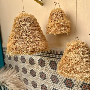 May include: Three hanging pendant lights with a natural, textured, straw-like material. The lampshades are bell-shaped and suspended from thin wires. The background features a patterned tile wall and a decorative metal bracket.
