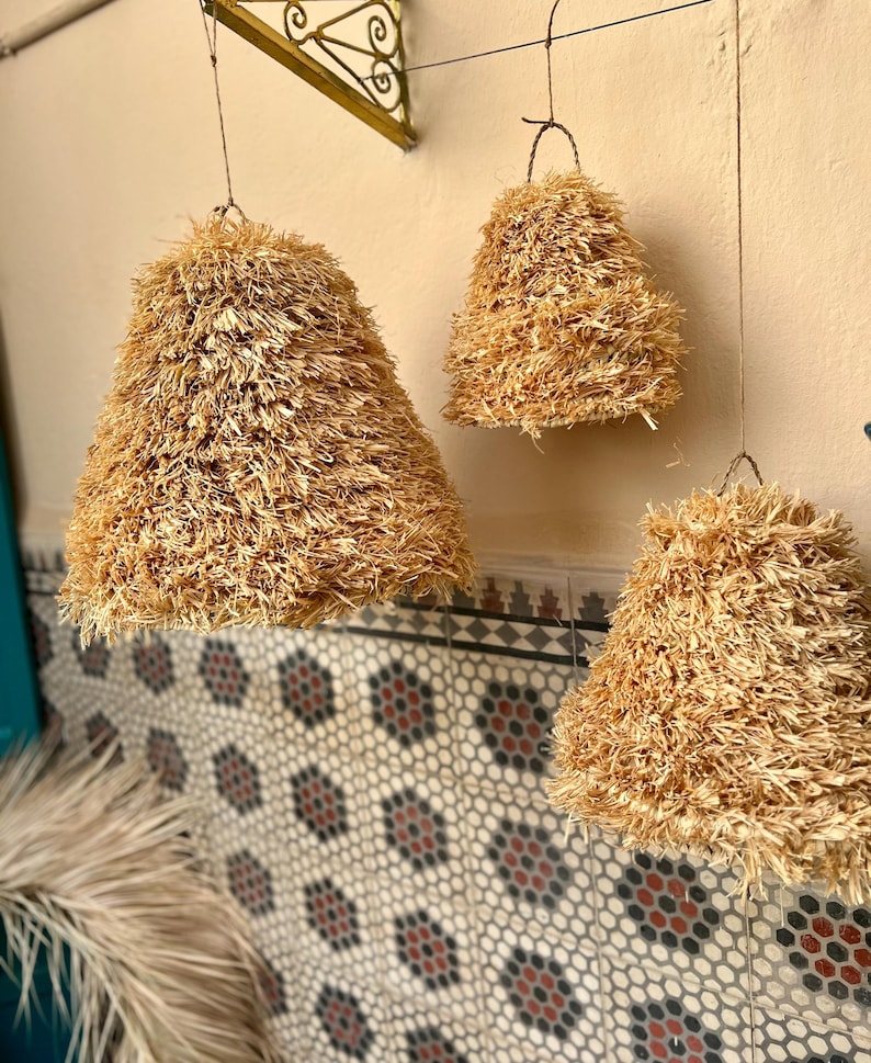 May include: Three hanging pendant lights with a natural, textured, straw-like material. The lampshades are bell-shaped and suspended from thin wires. The background features a patterned tile wall and a decorative metal bracket.