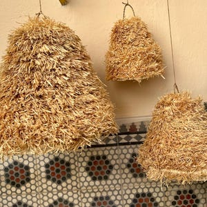 May include: Three hanging pendant lights made of natural straw. The lights are cone-shaped, with a textured, layered appearance. They are suspended from thin wires against a neutral-colored wall. The lights vary in size, creating a visually interesting display.