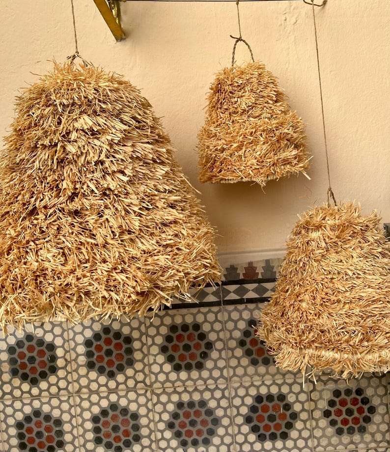 May include: Three hanging pendant lights made of natural straw. The lights are cone-shaped, with a textured, layered appearance. They are suspended from thin wires against a neutral-colored wall. The lights vary in size, creating a visually interesting display.