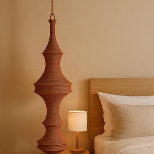 May include: A tall, rust-colored, crocheted pendant light fixture with a complex, multi-tiered, hourglass-like silhouette, suspended from the ceiling by a thick, braided rope cord, hanging above a small wooden side table.