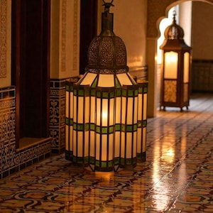 May include: A decorative hanging lantern with a bronze top and a stained-glass design. The lantern has white and green panels, casting a warm light. The floor is tiled with a detailed pattern, and another lantern is visible in the background, creating a warm ambiance.