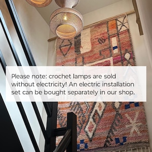 May include: Three woven pendant lamps with a light brown finish hanging above a large, rectangular wall tapestry featuring a multi-colored geometric pattern in shades of orange, pink, and blue with fringe at the bottom edge and black railing.