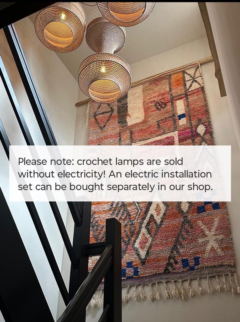 May include: Three woven pendant lamps with a light brown finish hanging above a large, rectangular wall tapestry featuring a multi-colored geometric pattern in shades of orange, pink, and blue with fringe at the bottom edge and black railing.