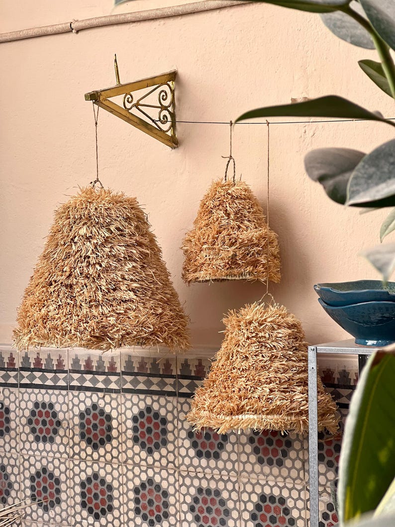 May include: Three straw lampshades of varying sizes hang from the ceiling. The lampshades are cone-shaped and made of a light brown, textured material. A decorative metal bracket is mounted on the wall. The background features patterned tiles and a glimpse of greenery.