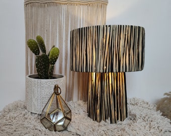 Puede incluir: Una lámpara de mesa a rayas blancas y negras con una pantalla de vidrio geométrica. La lámpara está sobre una alfombra blanca esponjosa con una planta de cactus en una maceta blanca en el fondo.