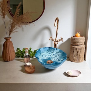 May include: A blue ceramic bathroom sink with a distinctive, ruffled edge. The sink features a textured, bubbly interior and is paired with a gold tap. Decorative items include a brown vase, a small pink plate, and a candle.