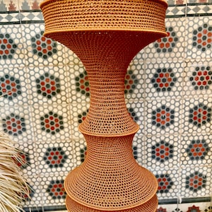 May include: A tall, hourglass-shaped pendant light fixture crafted from a rust-colored, tightly woven cord material. The structure features a wide, flared top section, a narrow central neck, and a tiered, rounded base with a consistent mesh texture.