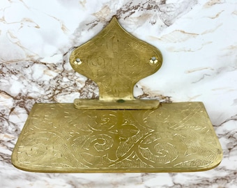 May include: A gold-colored brass shelf with intricate floral designs. The shelf is attached to a wall with a decorative, pointed backplate.