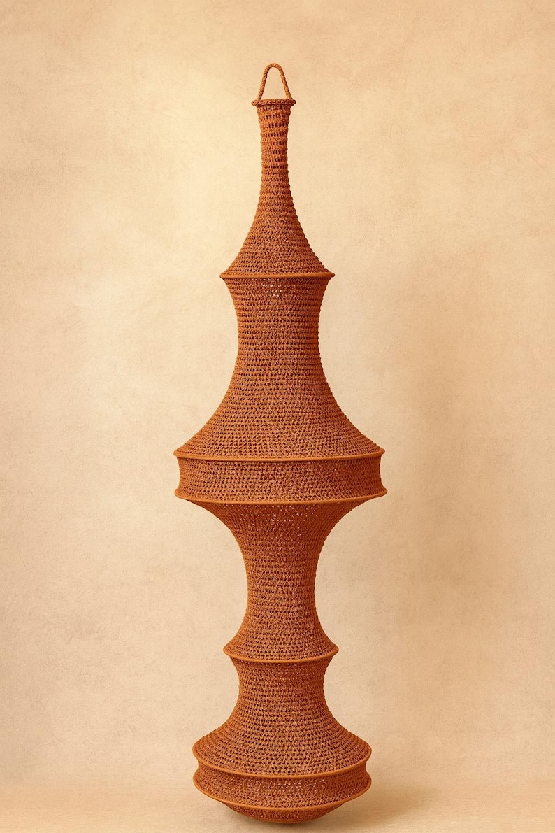 May include: A tall, rust-colored, crocheted hanging lamp shade with a complex, multi-tiered silhouette. The structure features a narrow neck, a wide central section, and a flared base, all crafted from a dense, uniform yarn stitch pattern.