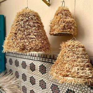 May include: Three hanging pendant lights with a natural, textured, straw-like material. The lampshades are cone-shaped and suspended from a wire. The background features a patterned tile wall and a decorative metal bracket.