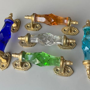 May include: Six decorative cabinet door pulls with faceted glass knobs in blue, orange, clear, green, and turquoise. Each knob is attached to a brass base with ornate detailing.