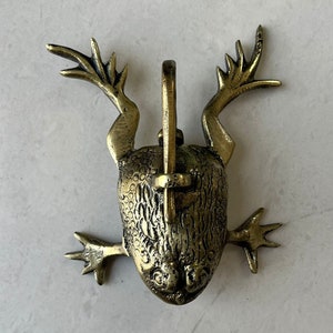 Puede incluir: Un llamador de puerta de color dorado en forma de rana con un diseño detallado. La rana tiene una lengua larga y las patas extendidas.