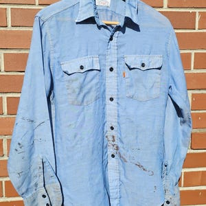 May include: Light blue button-down shirt with two chest pockets and black buttons. The shirt has long sleeves and a collar. There are stains on the shirt. The shirt is hanging on a wooden hanger.