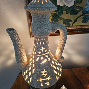 May include: White ceramic lamp shaped like a teapot with intricate cutouts and a wooden base. The lamp is lit and casting a warm glow. The lamp has Chinese characters on the side.