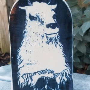 May include: A ceramic tile with a black and white image of a goat's head. The goat is facing to the right and has a white face with black eyes and a black nose. The background is black.