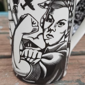May include: White ceramic mug featuring a black and white illustration of a woman flexing her bicep, reminiscent of the Rosie the Riveter image. The mug also has a black and white airplane and other design elements.