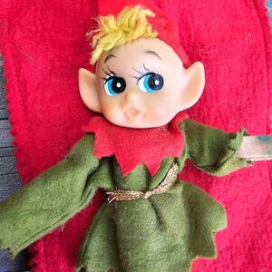 May include: A vintage elf doll with a green and red outfit, blonde hair, and large blue eyes. The elf is lying on a red felt surface.