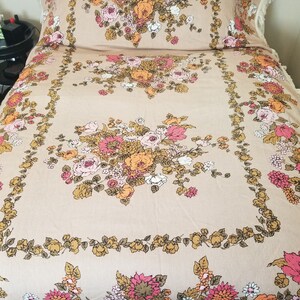May include: A vintage floral bedspread with a beige background and a large floral design in shades of pink, orange, and yellow. The bedspread has a fringe border.