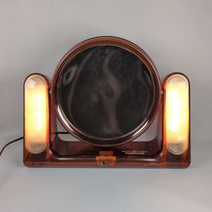 May include: A vintage brown vanity mirror with two side lights. The round mirror is framed in brown plastic and has a built-in on/off switch. The lights are rectangular with a spiral design and emit a warm glow.
