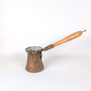 May include: A copper coffee pot with a wooden handle. The pot has a rounded base and a narrow spout. The handle is light brown and attached to the pot with a metal piece. The pot is designed for brewing coffee.