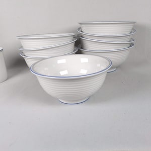 May include: A collection of white ceramic bowls with a ribbed texture and a thin blue trim. The bowls are stacked and arranged, showcasing their design. The image highlights the clean lines and simple elegance of the tableware.