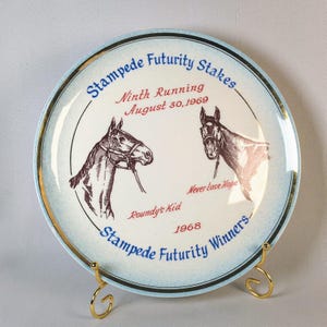 May include: A decorative plate with a light blue rim and gold trim, featuring two horse head illustrations. The plate commemorates the Stampede Futurity Stakes, with text indicating the ninth running on August 30, 1969, and the names "Roundy's Kid" and "Never Lose Hope."
