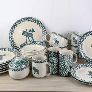 Può includere: Una collezione di stoviglie in ceramica color crema con un design a alce blu. Il set include piatti, ciotole, tazze, una lattiera e una zuccheriera. I bordi presentano un motivo blu a pois. I piatti hanno un diametro di circa 25 cm.