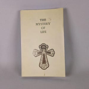 May include: A vintage book with a cream-colored cover. The title "THE MYSTERY OF LIFE" is printed in black, above a decorative cross illustration. The cross is detailed with floral and ornate patterns.
