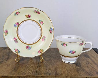 May include: A vintage teacup and saucer set. The pale yellow cup and saucer feature a floral pattern with pink and blue flowers and gold trim. The saucer is on a gold stand.